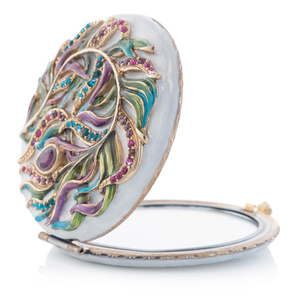 Rowan Classic Peacock Compact. White enamel compact mirror with an enameled peacock feather adorned with purple, pink, and blue crystals. View of left side, opened.