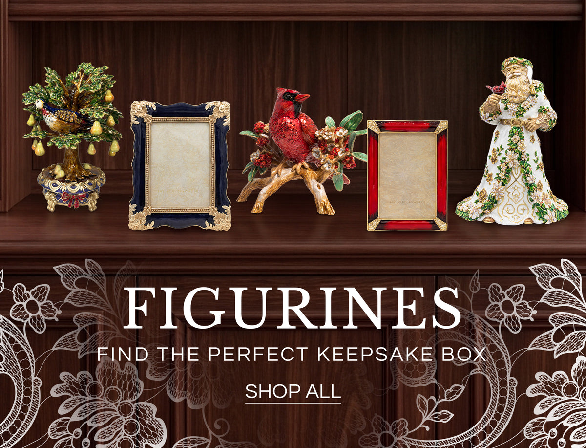 Promotional banner for "FIGURINES - FIND YOUR NEXT TREASURE" featuring Jay Strongwater decor pieces arranged on a dark wood mantle: a jeweled partridge pear tree figurine, a dark blue enamel photo frame with ornate gold trim, a red cardinal perched on a leafy branch figurine, a red crystal picture frame, and an elaborate white-and-gold Santa figurine holding a small bird.
