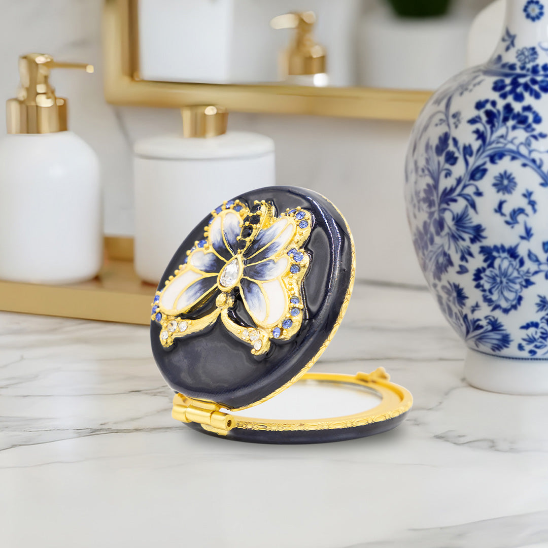 Navy compact mirror with a blue butterfuly on the outside sitting on a bathroom counter