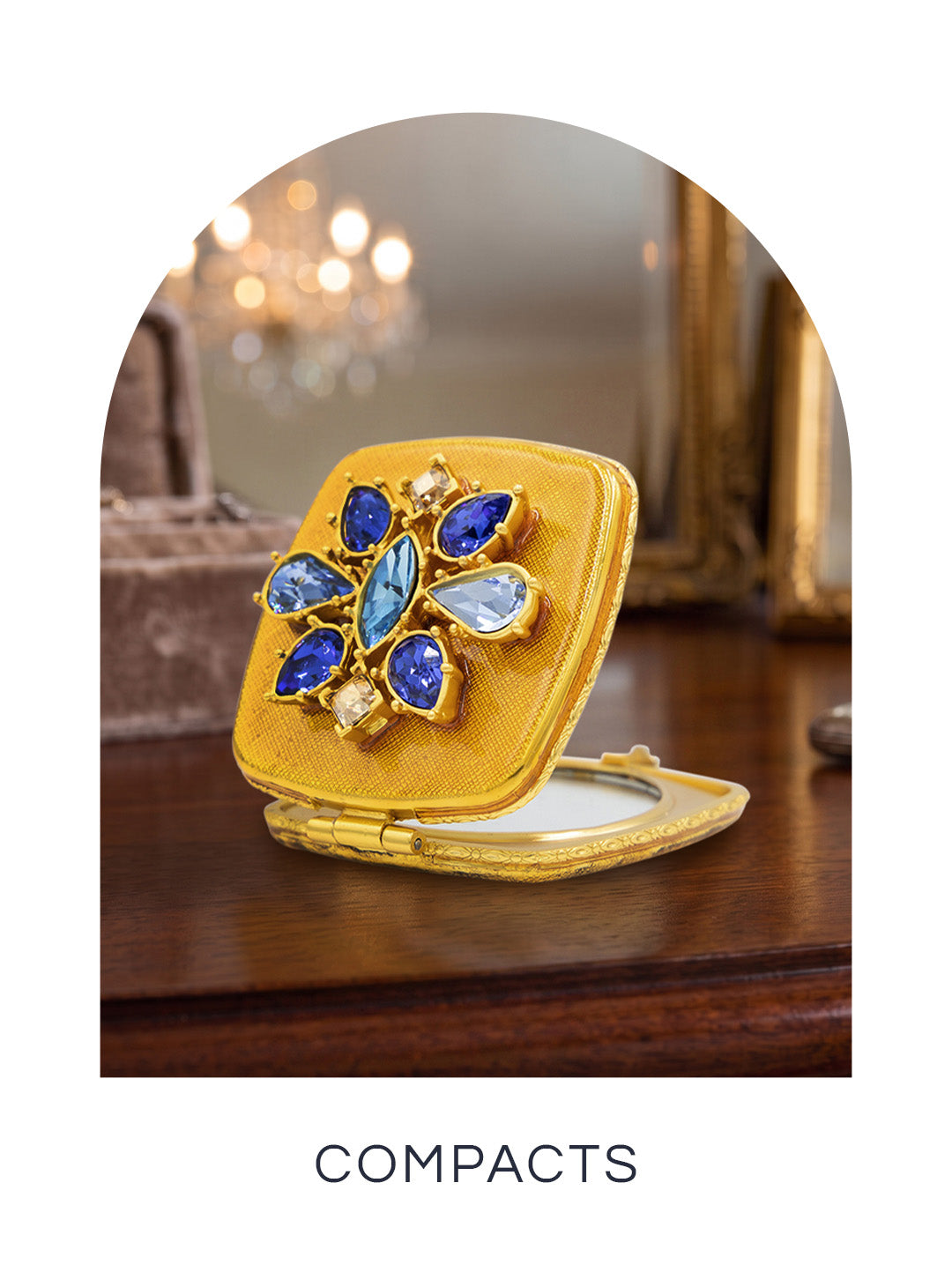 Jeweled gold compact mirror adorned with a floral motif of blue and light blue crystals, displayed partially open on a wooden vanity for the Compacts collection.